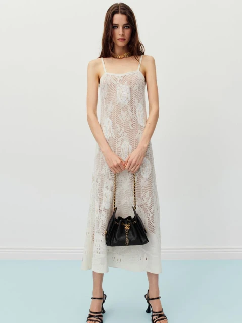 A full shot of a female fashion model showcasing a semi-sheer, rose-patterned dress, a black purse, and black high-heel shoes in a studio setting.