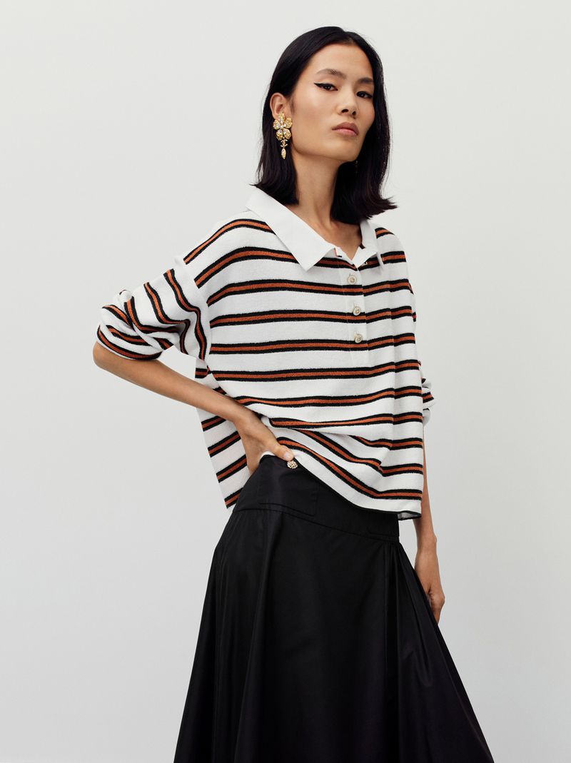 An Asian woman poses in a studio, wearing a striped knit top with a white collar, gold statement earrings, and a black skirt.