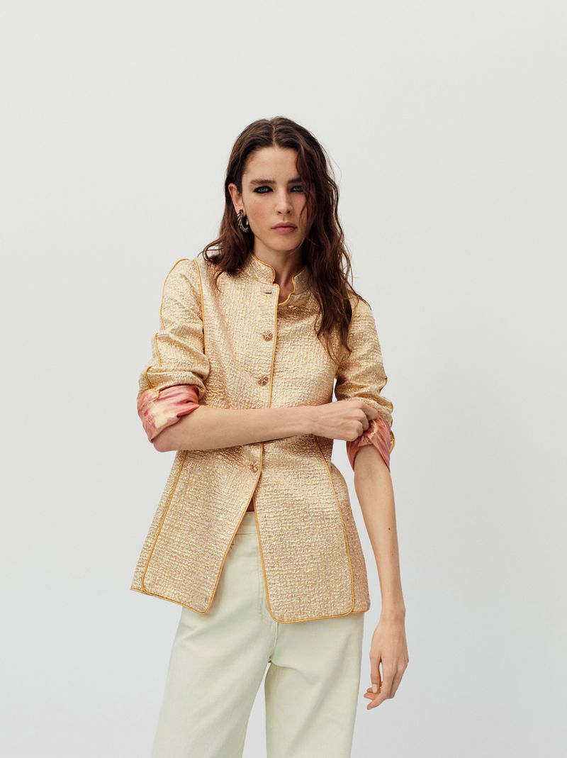 A fashionable adult woman is posing indoors wearing a gold jacket with pale yellow pants. The background is a plain white wall.