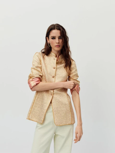 A fashionable adult woman is posing indoors wearing a gold jacket with pale yellow pants. The background is a plain white wall.