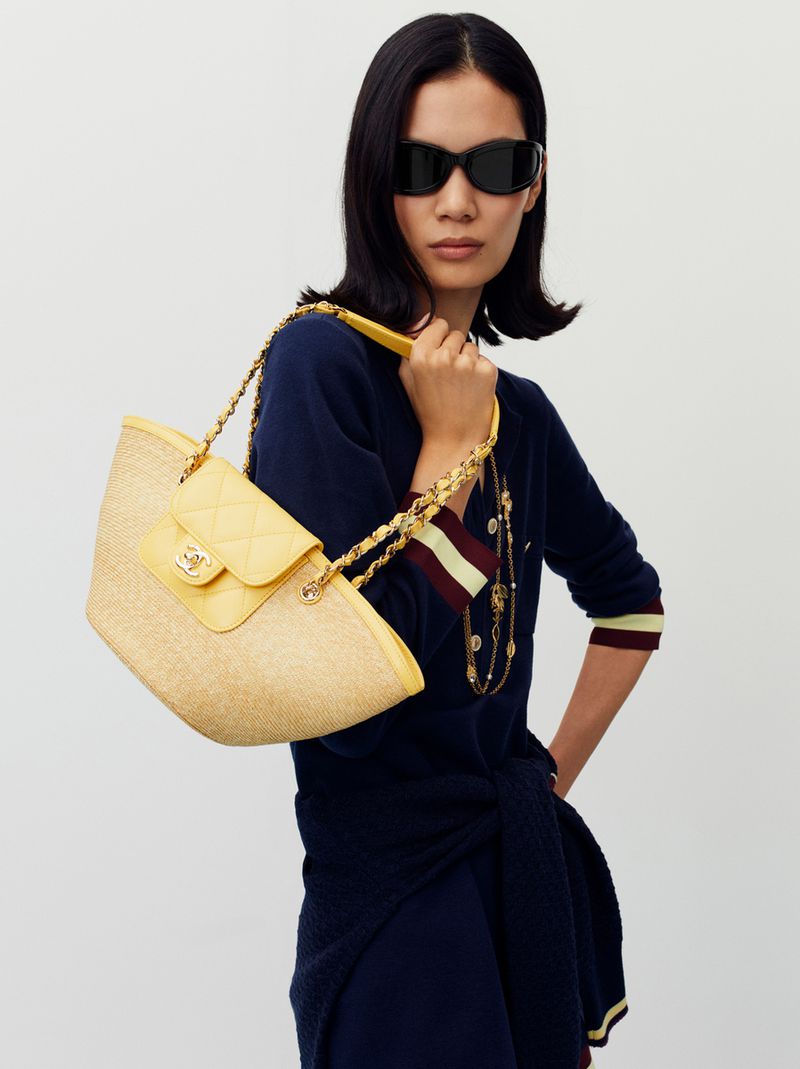Asian woman models straw Chanel handbag with yellow leather flap and gold chain strap, accessorized with sunglasses and dark sweater.