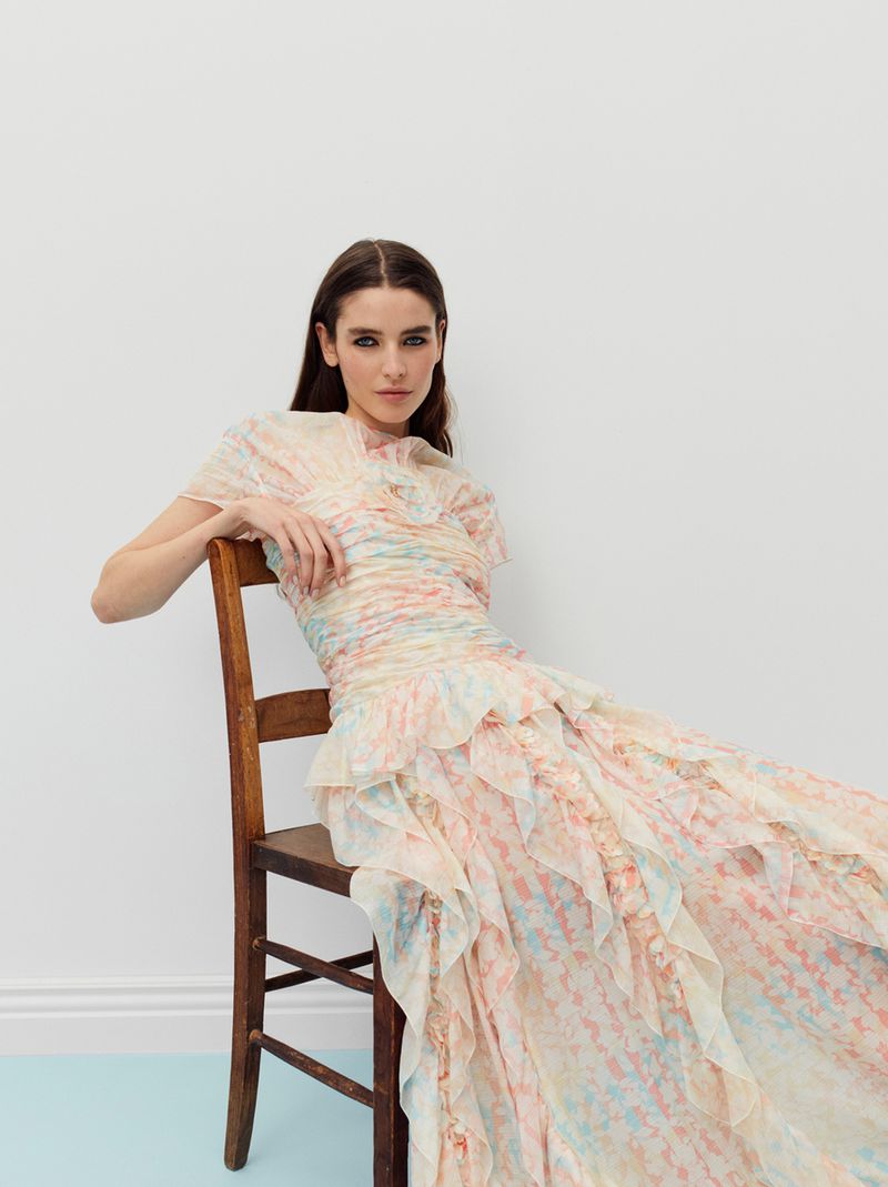 A fashionable adult female model with long brown hair sits on a wooden chair wearing a colorful, ruffled chiffon gown in a studio setting.