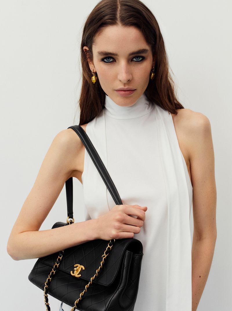 Elegant woman with long brunette hair and gold earrings holds a black Chanel shoulder bag against a plain white background.