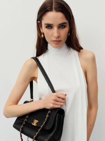 Elegant woman with long brunette hair and gold earrings holds a black Chanel shoulder bag against a plain white background.