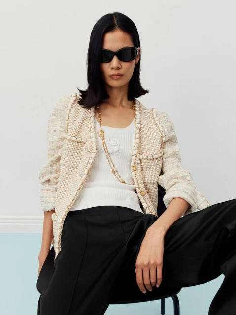 Woman in a bouclé jacket, sunglasses, and black trousers sits in a light studio. Fashion editorial shot with neutral background.
