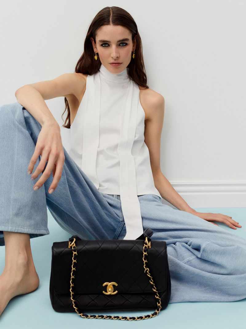 Fashion shot of a brunette woman in a white top and blue jeans, seated, with a black Chanel handbag with a gold chain.