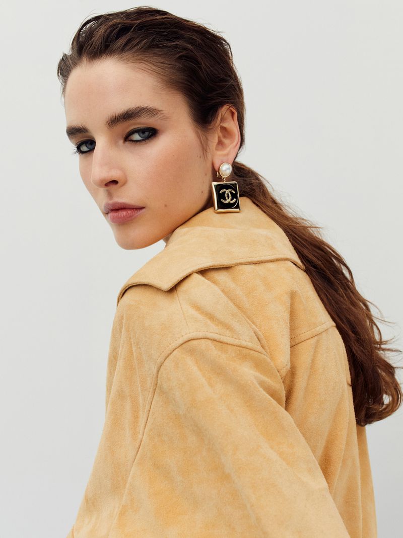 Portrait of a brunette woman with blue eyes wearing a light tan jacket and a pair of square Chanel logo pearl earrings.