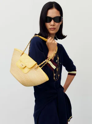 Model wearing dark sunglasses and a navy blue top carrying a yellow straw handbag against a plain white background