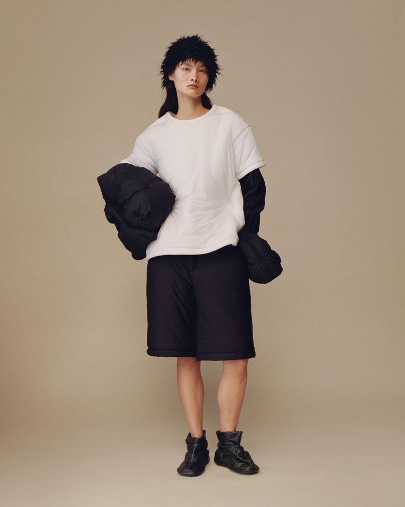 An Asian woman stands in a studio wearing a white top, black shorts, and black boots, holding a black jacket in front of a neutral background.