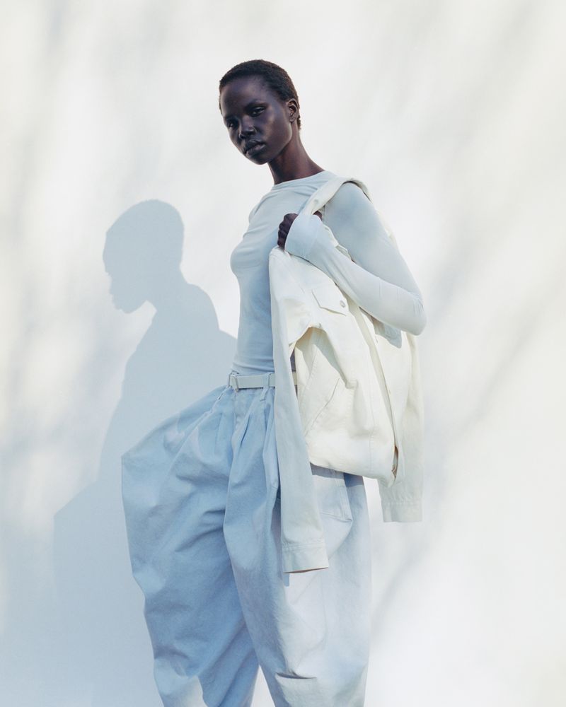 Woman wearing a long-sleeve shirt and loose pants with an off-white jacket draped over her shoulder against a bright white background.