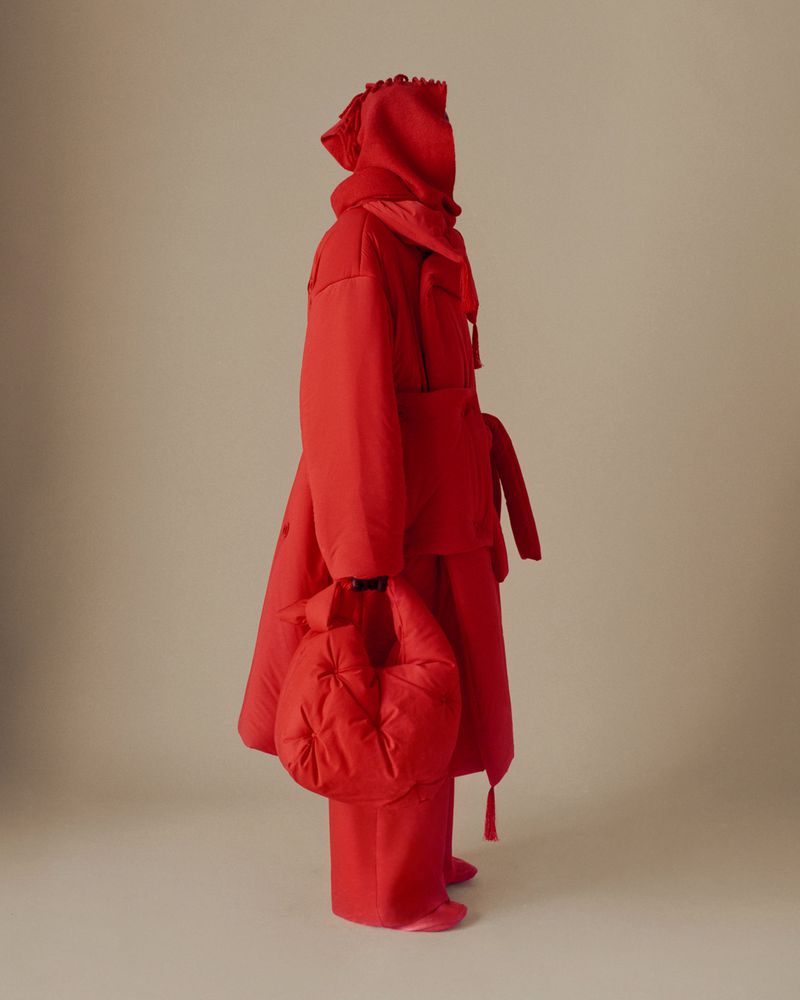 Fashion shot of a person wearing a full red monochromatic outfit with a hooded coat, trousers, and puffy bag, studio setting.
