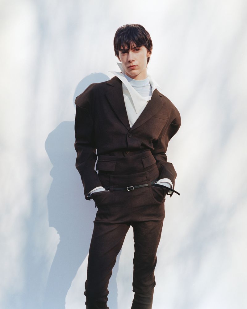 Person in a dark brown suit with an unusual silhouette, pale blue shirt, and white scarf, posing against a white background with shadows.