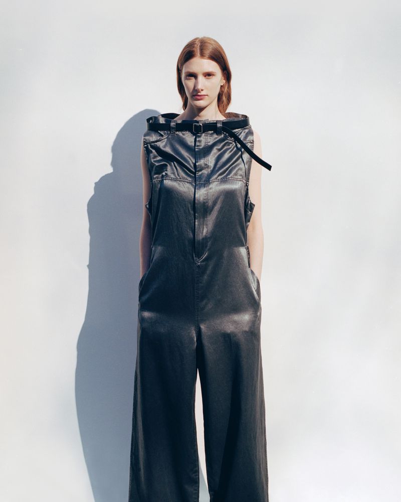 Fashion shot of a red-haired model in a shiny, dark grey sleeveless hooded jumpsuit, standing in a photo studio with soft lighting and shadows.