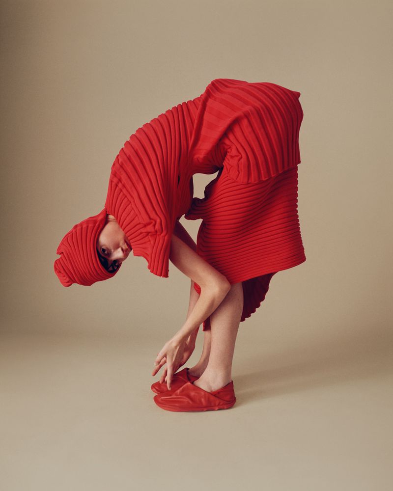 A woman in a red, pleated, hooded outfit is bent over with hands on floor in a studio setting, showcasing fashion and photography.
