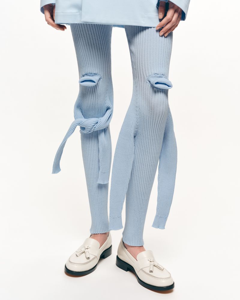 Closeup of a person wearing baby blue deconstructed ribbed knit leggings and white leather loafers on a clean white backdrop.