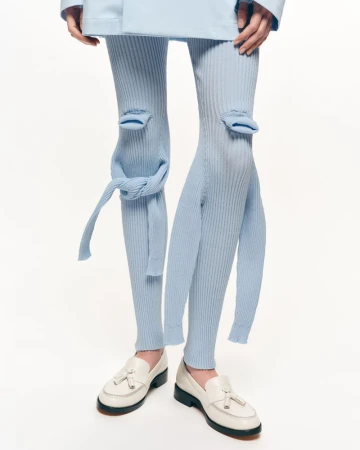 Closeup of a person wearing baby blue deconstructed ribbed knit leggings and white leather loafers on a clean white backdrop.