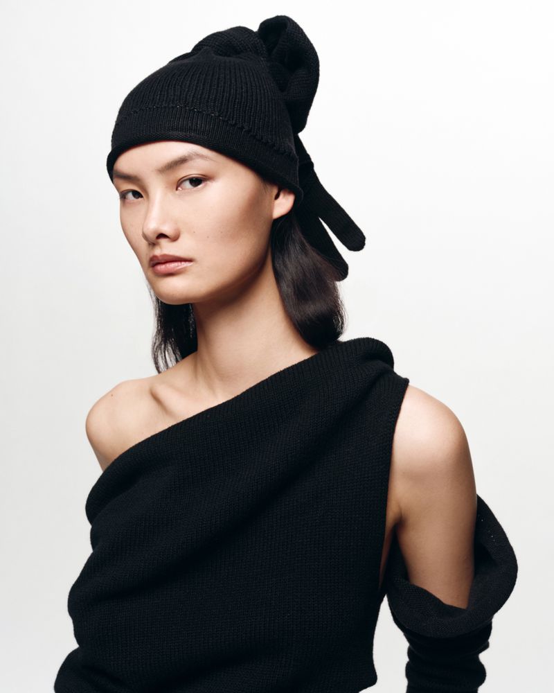 Portrait of a woman with an Asian appearance wearing a black knit hat and off-the-shoulder black sweater in a white studio setting.