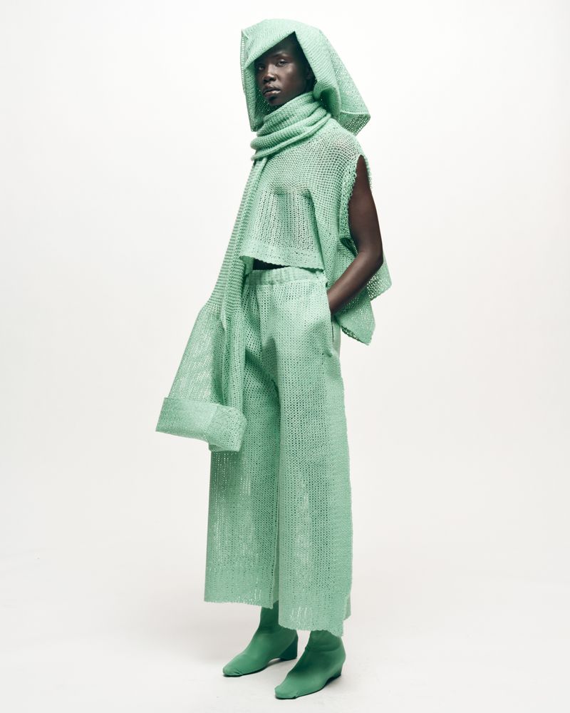 Fashion shoot: Black female model poses in a light green knit hooded top, scarf, pants, and booties against a clean white backdrop.