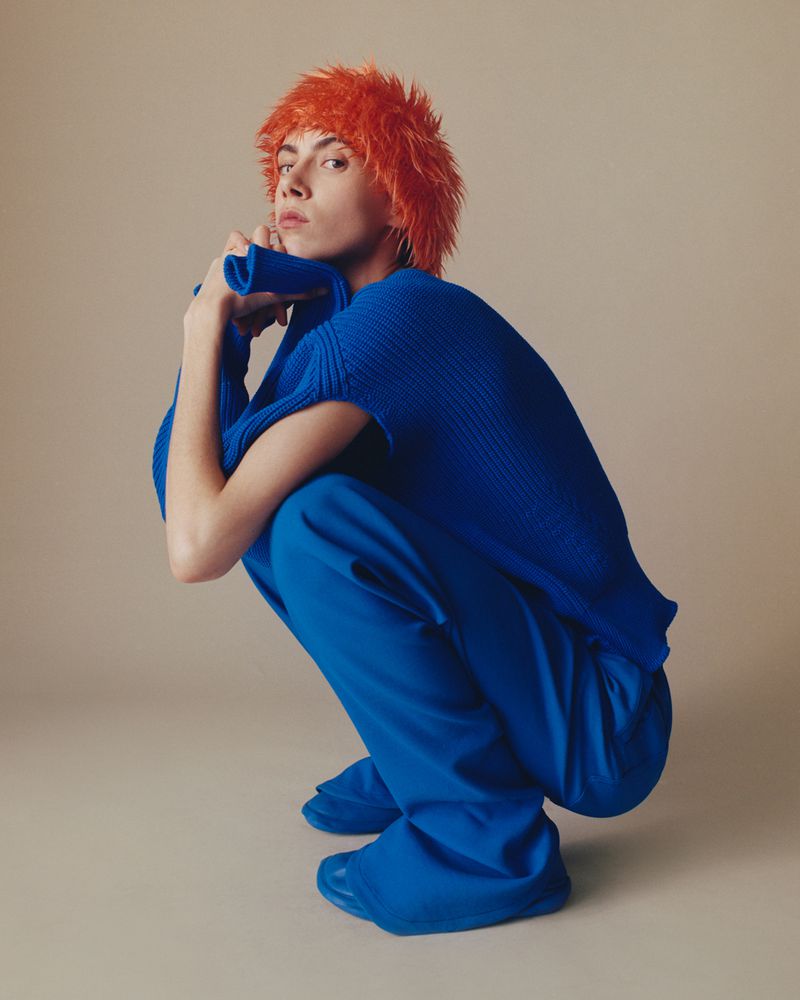 Person with bright orange hair and blue sweater and pants crouching in front of a neutral background in a fashion editorial shot.