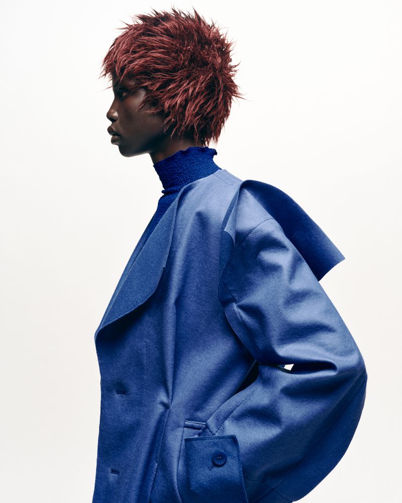 Profile fashion portrait of a Black woman with red hair wearing a blue coat against a white background in a photo studio.