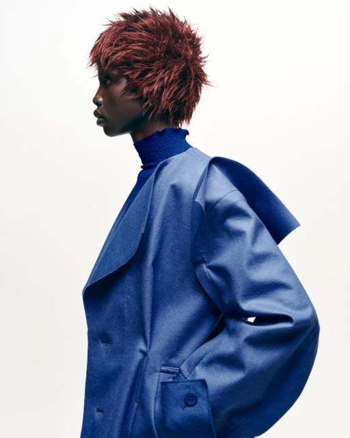 Profile fashion portrait of a Black woman with red hair wearing a blue coat against a white background in a photo studio.