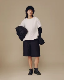 Model standing in a white short-sleeve top, black shorts, and black ankle boots against a beige studio background.