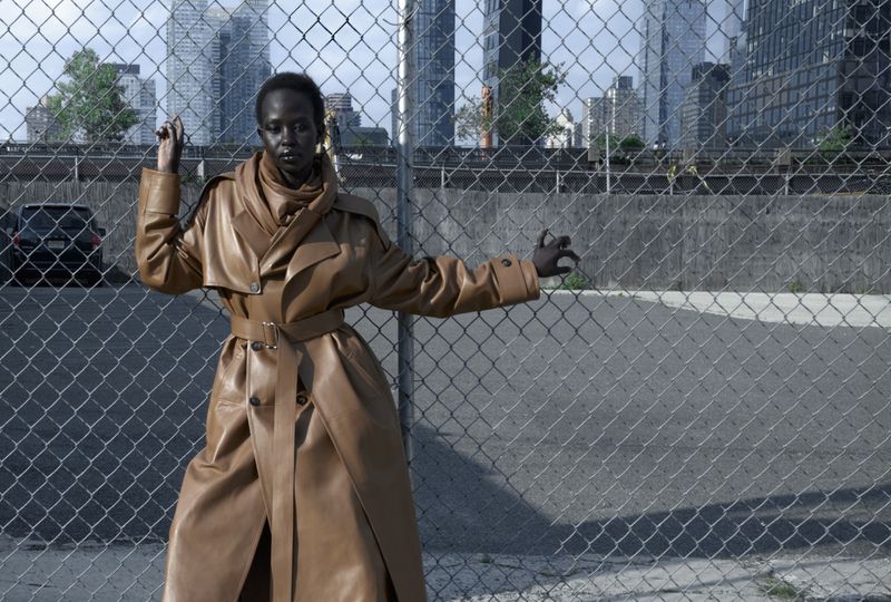 Woman poses in tan leather trench coat in front of chain-link fence with city buildings in the distance on an overcast day.