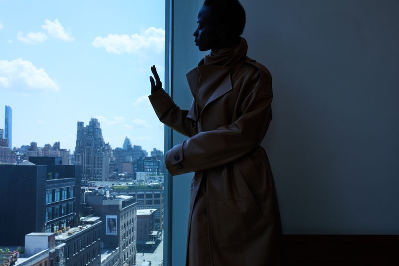 Woman in a tan trench coat looks out of a window at the city skyline. The image is split between light and shadow in an urban setting.