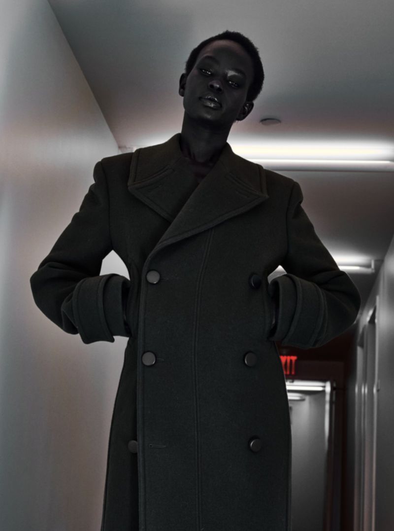 A striking portrait of an adult, dark-skinned model in a hallway wearing a structured black overcoat, emphasizing fashion and style.