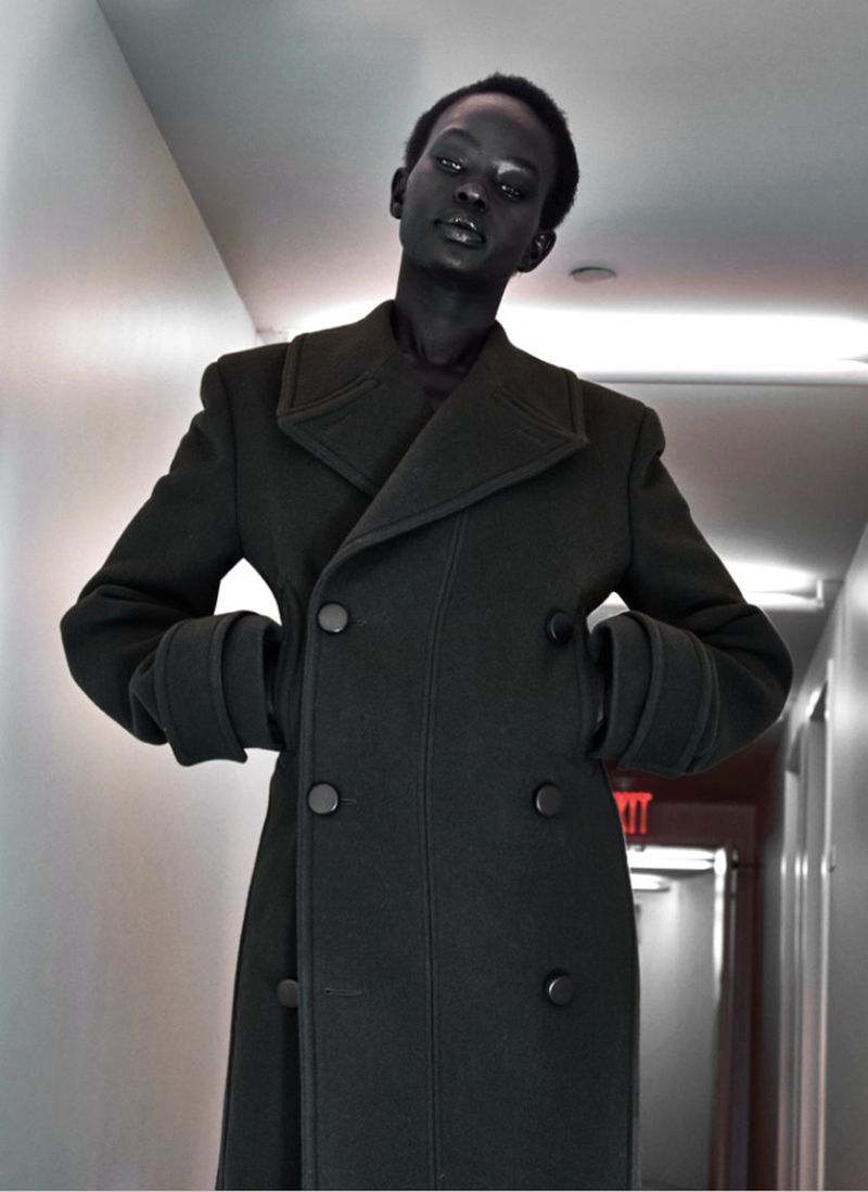 Person in an olive double-breasted wool coat stands in a hallway with hands in the pockets looking at the camera.