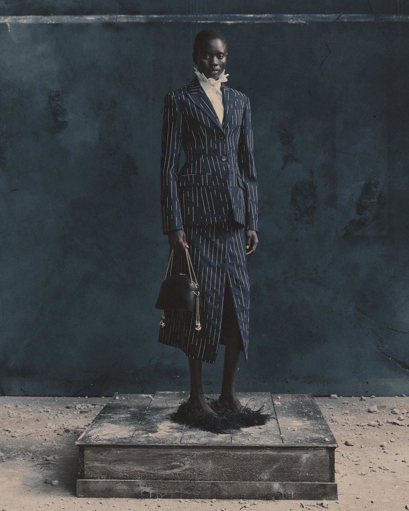 A slender Black woman in a pinstripe skirt suit and furry shoes poses on a stone platform against a dark, mottled wall.