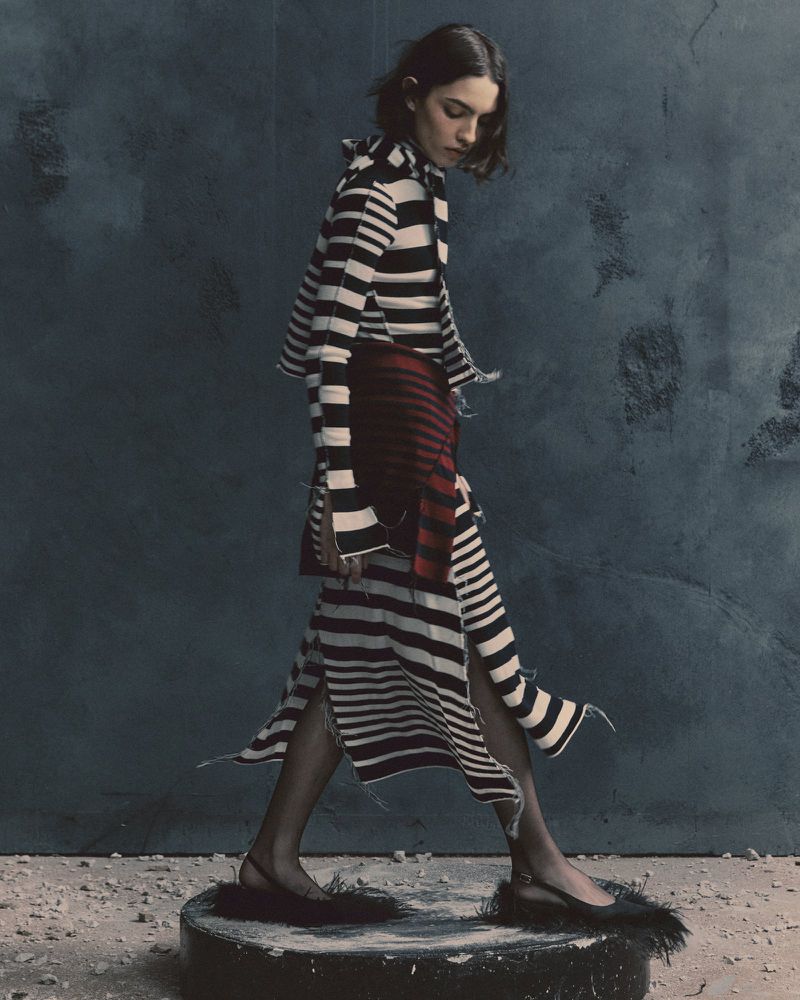 A female fashion model showcases a striped dress, accessorized with slingback flats featuring feather details, posed against a textured backdrop.