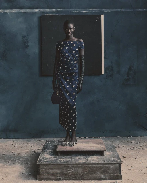 A slender woman with dark skin stands on a platform wearing a blue floral dress, long gloves, and bejeweled shoes in a studio setting.