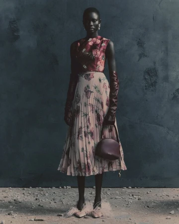 Model wearing a floral print sleeveless top and pleated skirt with long burgundy leather gloves standing against a dark wall