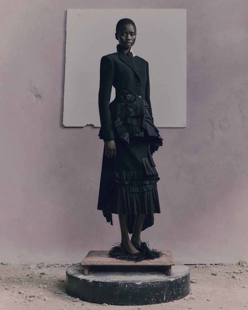 A fashion-style studio shot featuring a Black woman in a dark ruffled dress and coat, standing on a platform with a backdrop.