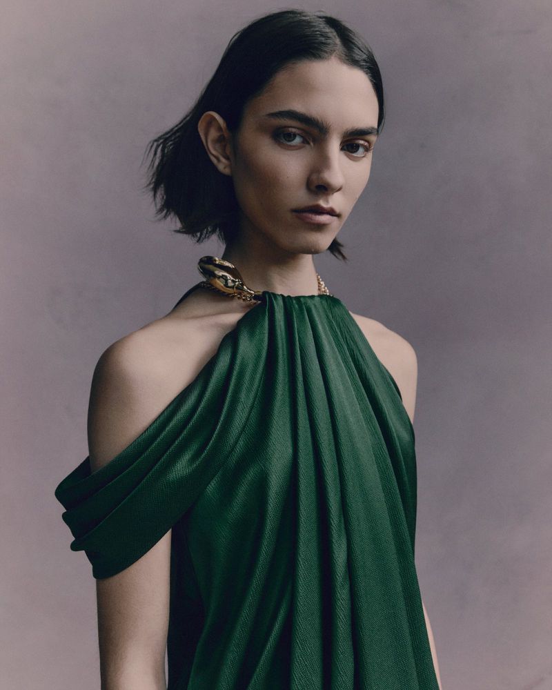 Woman wearing a green draped halter dress with a gold snake clasp poses in a studio with soft lighting and a neutral background.