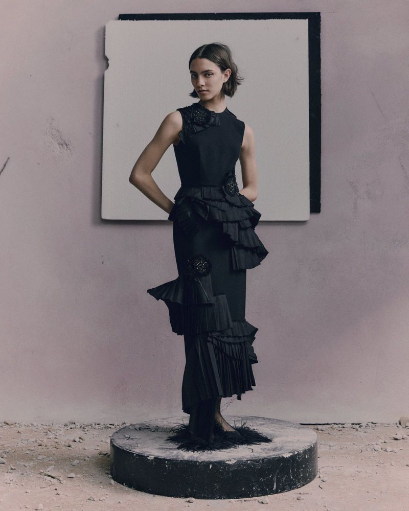 Woman in black sleeveless ruffled dress posing on a pedestal against a neutral backdrop, fashion editorial photography.