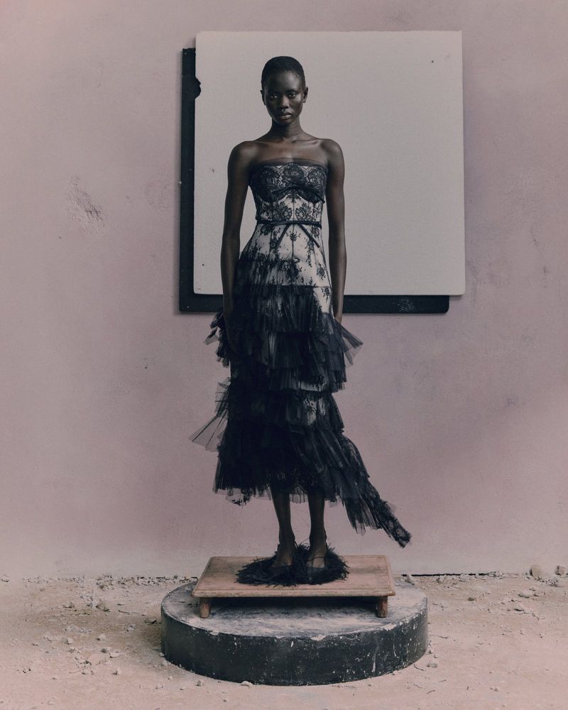 A solitary, stylish, African woman stands tall in a black strapless tiered dress, posing on a platform with an off-white background.
