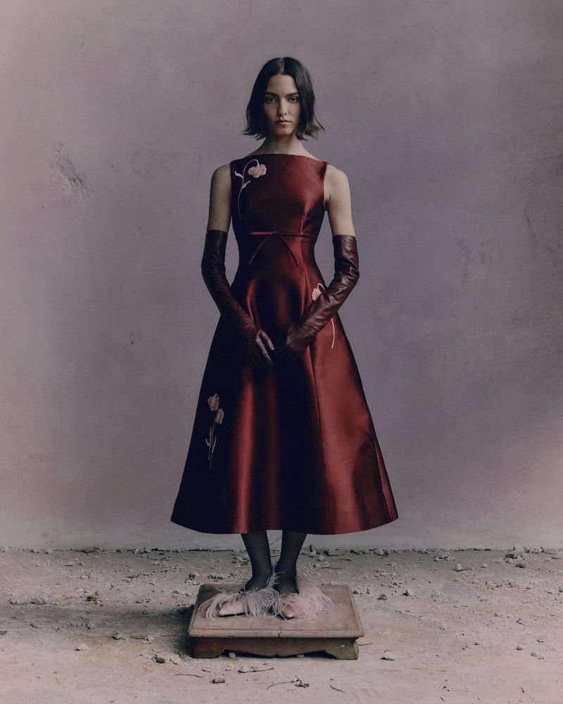 A woman in a red dress and opera gloves stands on a wooden platform in a neutral setting, fashion editorial shot, eye-level angle.