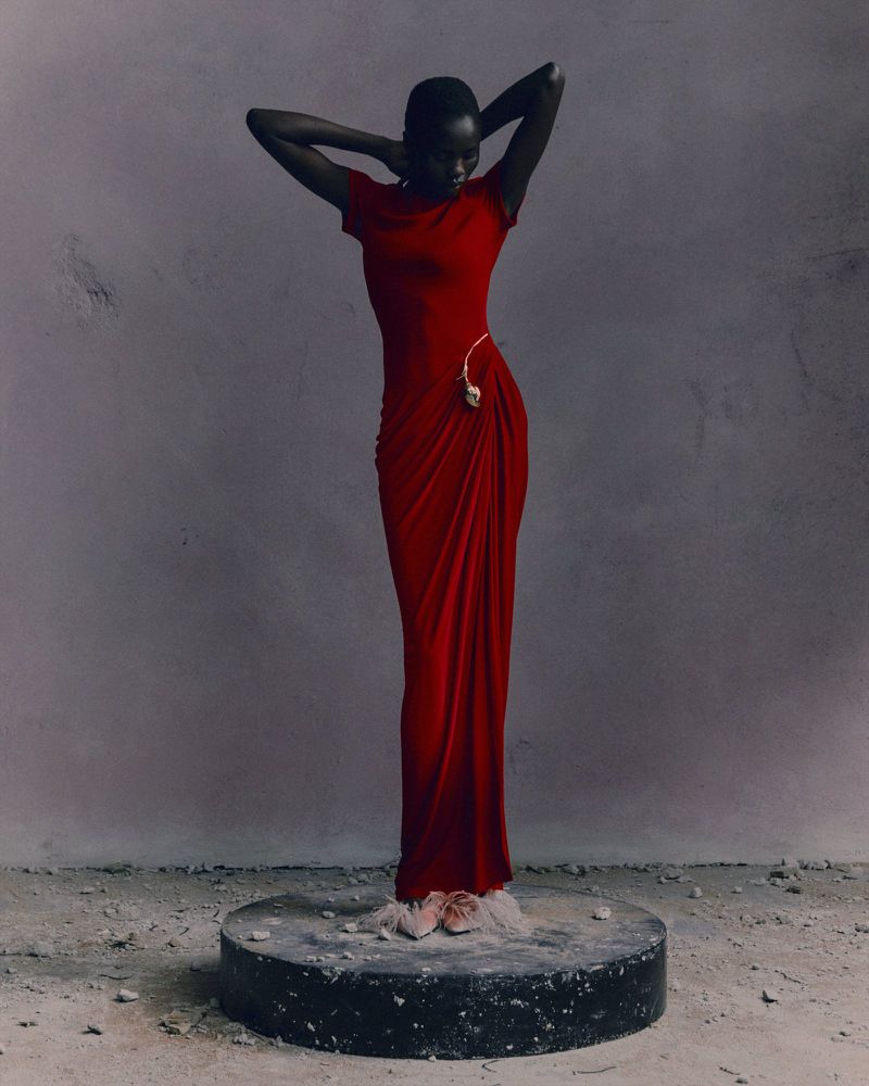 Elegant dark-skinned woman in a red gown stands on a round stage against a mottled gray background in a studio shot, fashion forward.