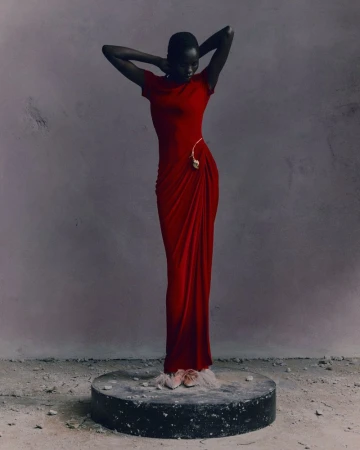Elegant dark-skinned woman in a red gown stands on a round stage against a mottled gray background in a studio shot, fashion forward.