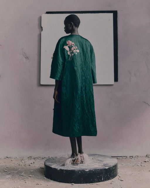 A black adult woman wearing a dark green coat with floral accents, standing on a round platform in front of a muted lavender wall.