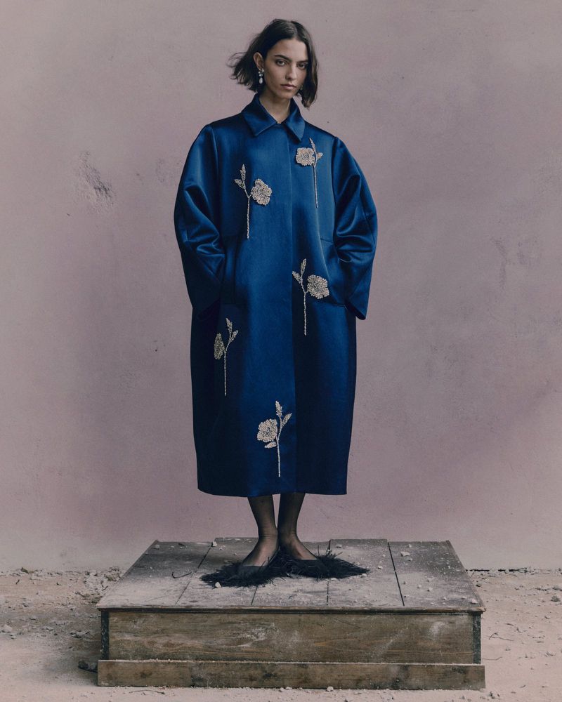 A young woman poses in a luxurious blue embroidered coat, standing on a concrete platform against a mottled mauve-colored backdrop.