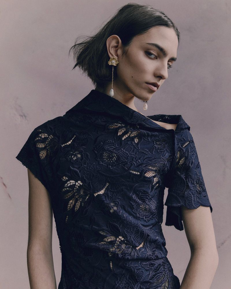 Elegant adult female poses in a studio setting, wearing a blue floral lace top and pearl earrings, showcasing fashion and beauty.