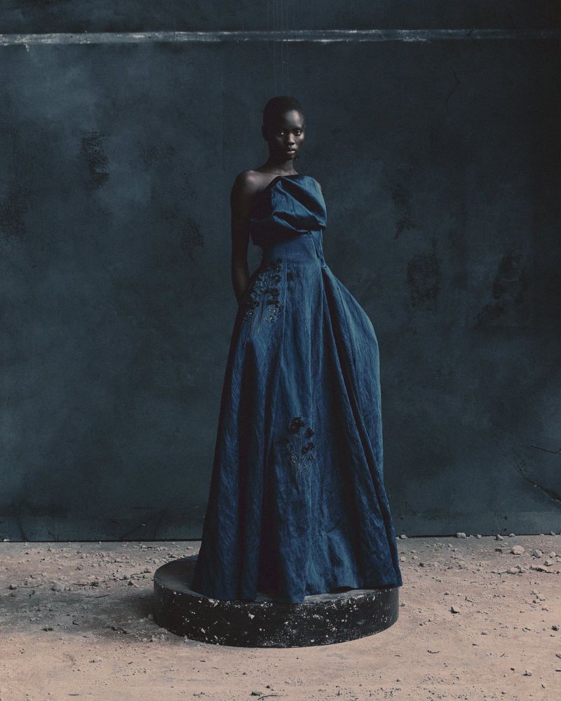 Elegant, dark-skinned woman stands in a draped blue gown, hands behind her back, on a concrete platform against a textured blue backdrop.