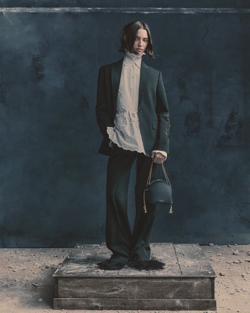A person in a dark suit and white shirt stands on a platform holding a dark handbag in a fashion editorial shot against a dark backdrop.