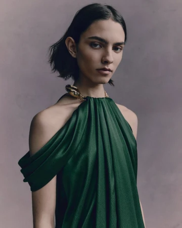 Model wearing a dark green pleated halter dress with gold collar detail against a muted grey studio background.