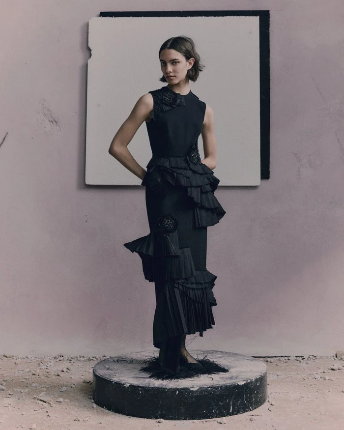 Model posing in a tiered black sleeveless ruffle dress on a circular stone plinth in a minimalist set with a textured backdrop.