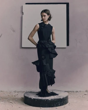 Model posing in a tiered black sleeveless ruffle dress on a circular stone plinth in a minimalist set with a textured backdrop.