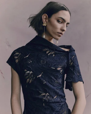Model in an asymmetrical navy embroidered lace top and drop pearl earrings against a muted textured backdrop.